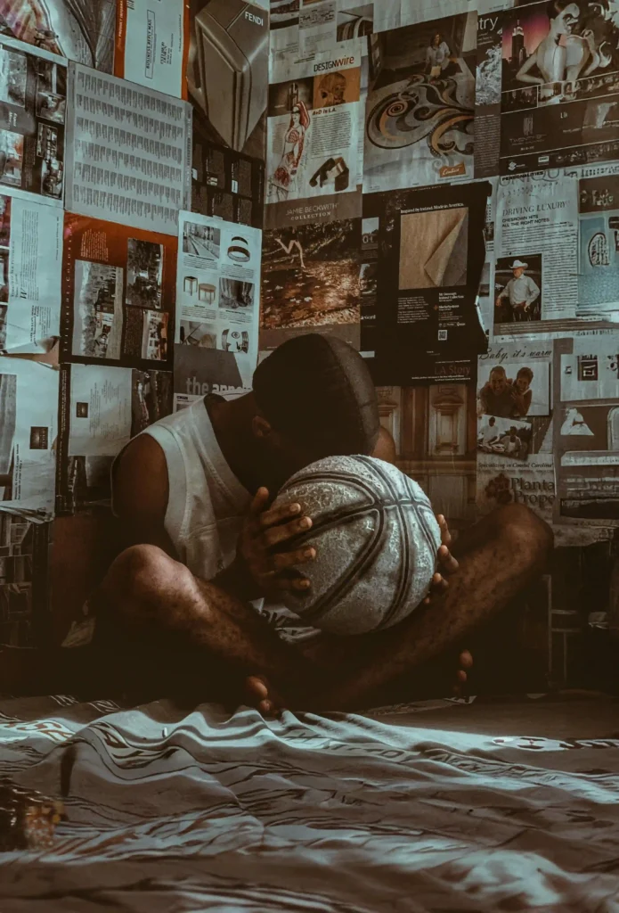 "A reflective basketball player sitting in a room filled with magazine clippings, holding a worn basketball."