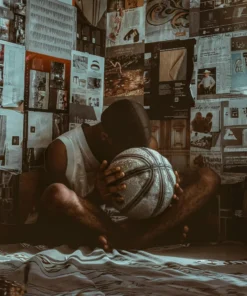 "A reflective basketball player sitting in a room filled with magazine clippings, holding a worn basketball."